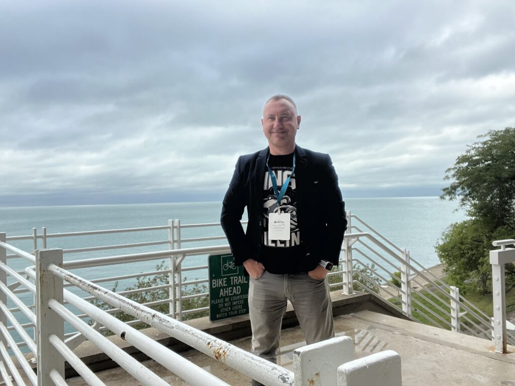 A man stands smiling on a balcony overlooking the ocean.