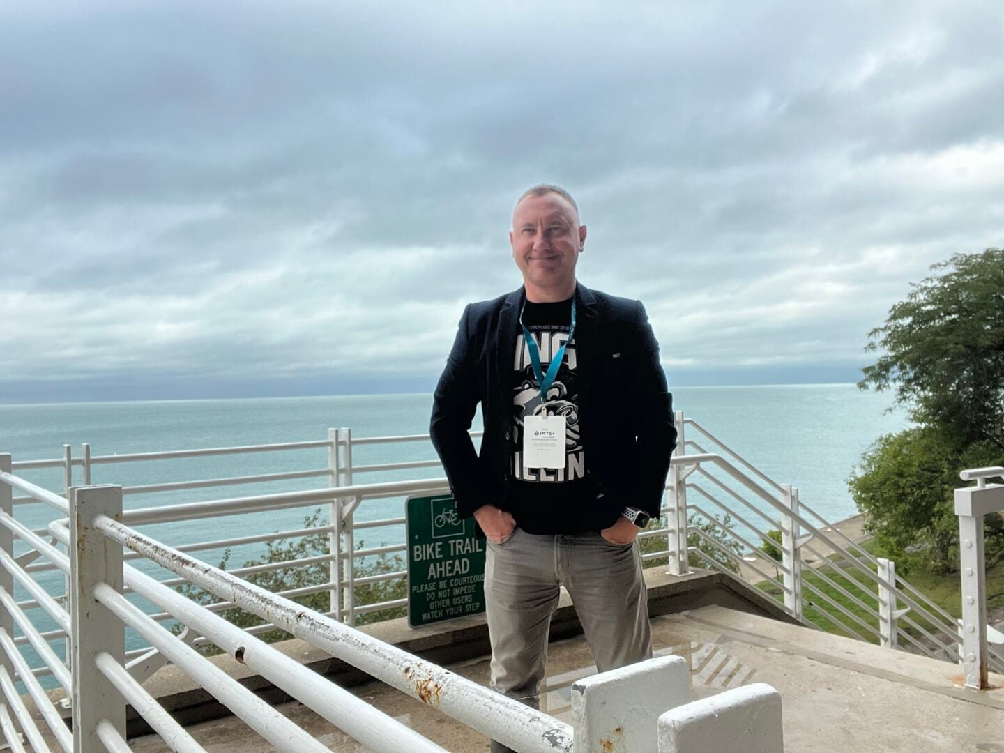 A man stands smiling on a balcony overlooking the ocean.