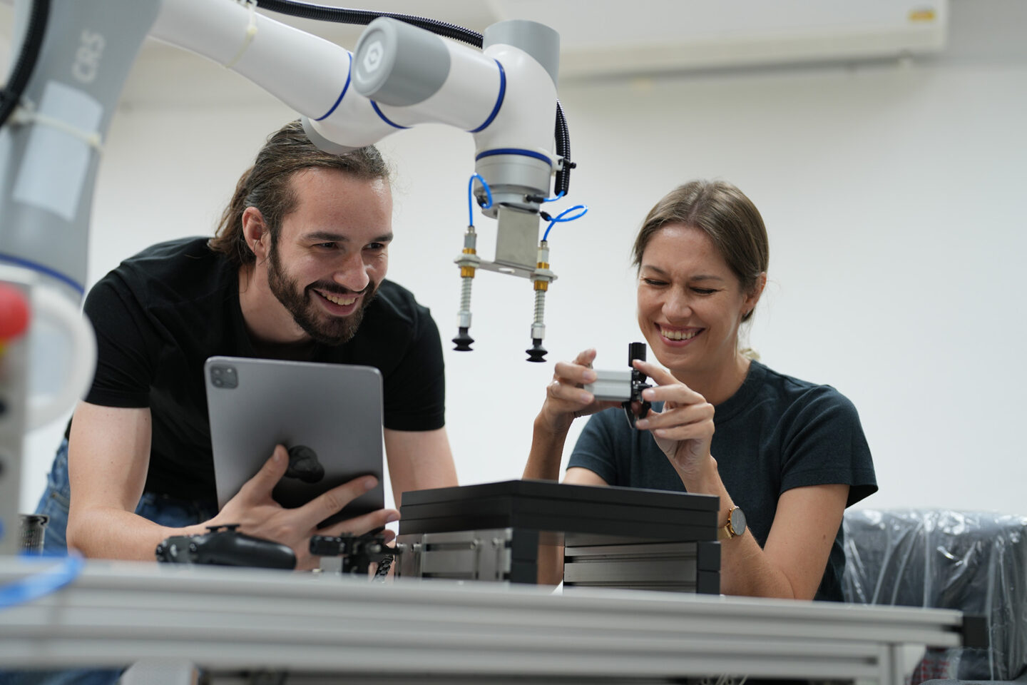 A man and woman observe a robotic arm while analyzing data on devices.