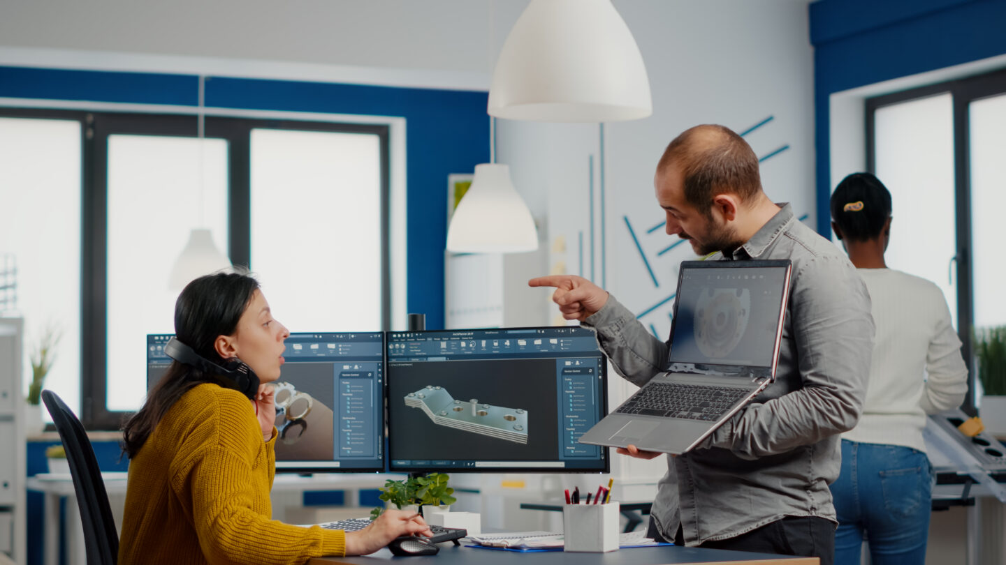 A project supervisor holds a laptop while pointing at a colleague.