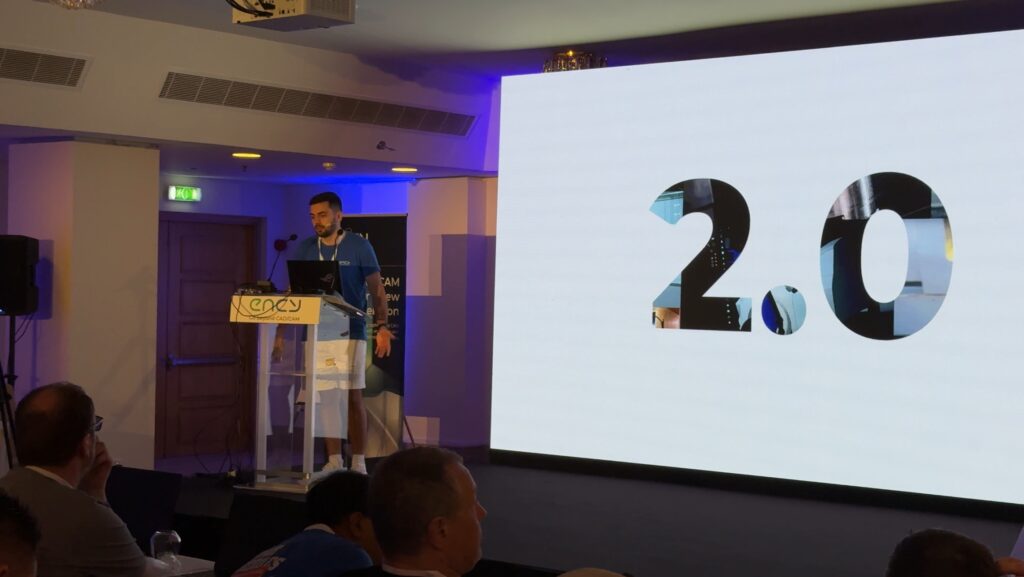 A presenter stands at a podium while showcasing a large number two on a screen.