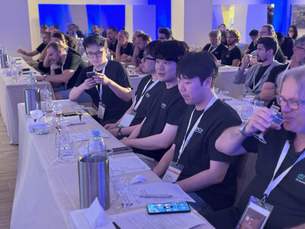 A group of attendees in black shirts listens intently at a conference.