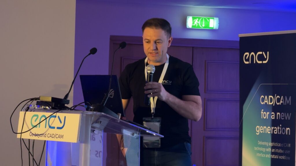 A speaker presents at a conference, standing beside a laptop.
