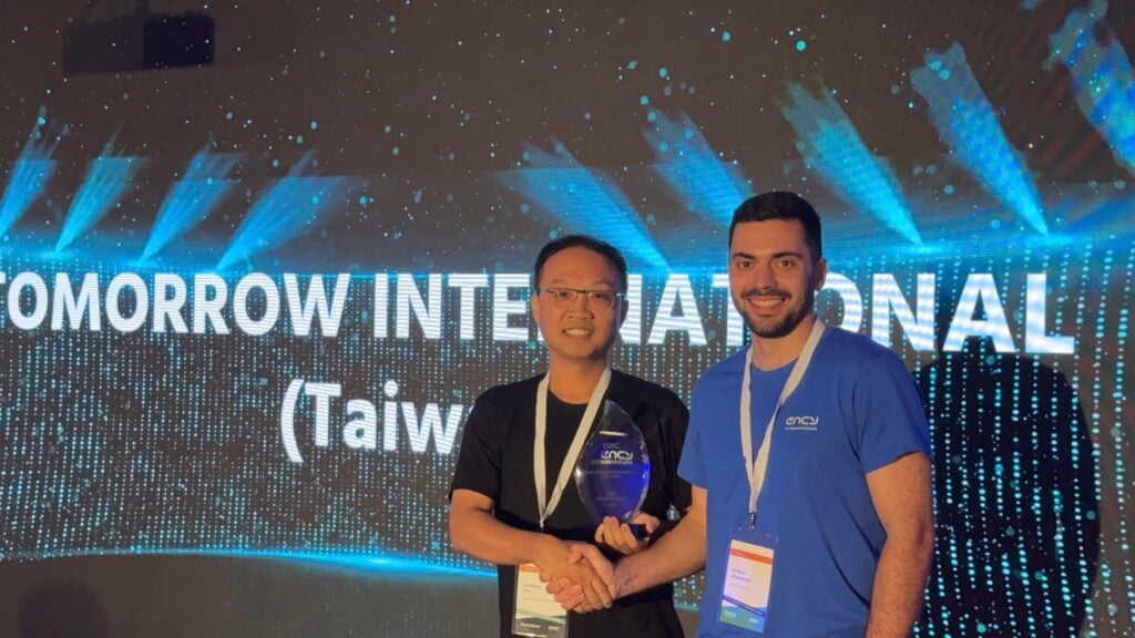 Two men shake hands on stage, holding an award together.