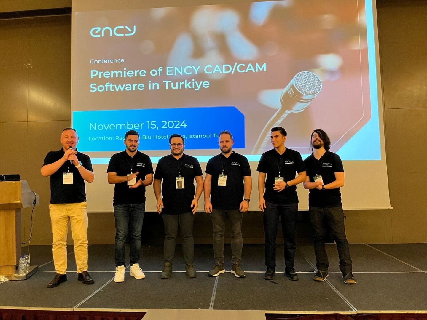 Six men in matching black shirts stand at a conference presentation.