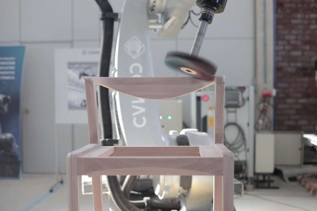 A robotic arm prepares to polish a wooden chair in a workshop.