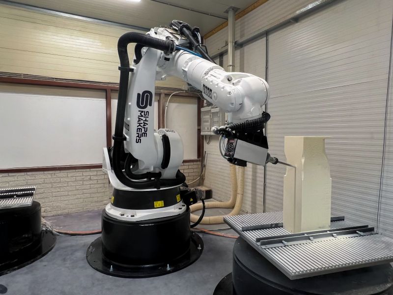 Six-axis industrial robot arm milling a light-colored foam or composite block into a contoured shape in an automated machining cell