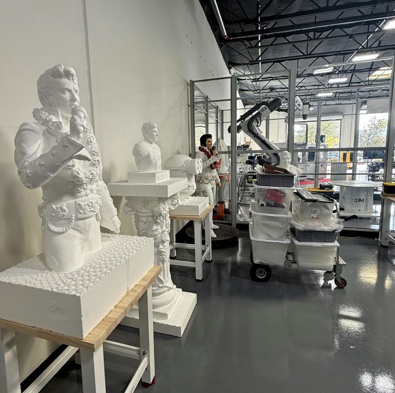 White six-axis industrial robot beside octagonal rotary table holding metal test coupons, with safety panels enclosing the cell