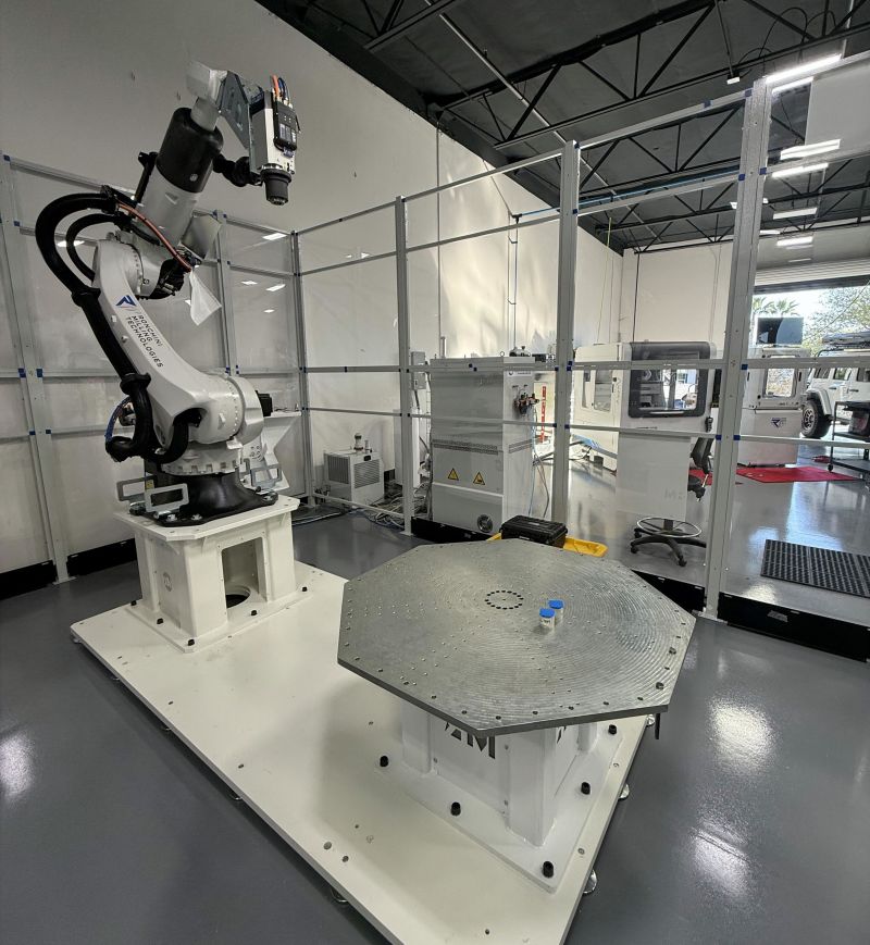 White six-axis industrial robot beside octagonal rotary table holding metal test coupons, with safety panels enclosing the cell