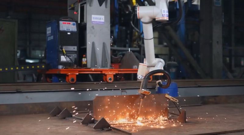 Six-axis collaborative robot arm performing automated welding on steel assembly with sparks flying
