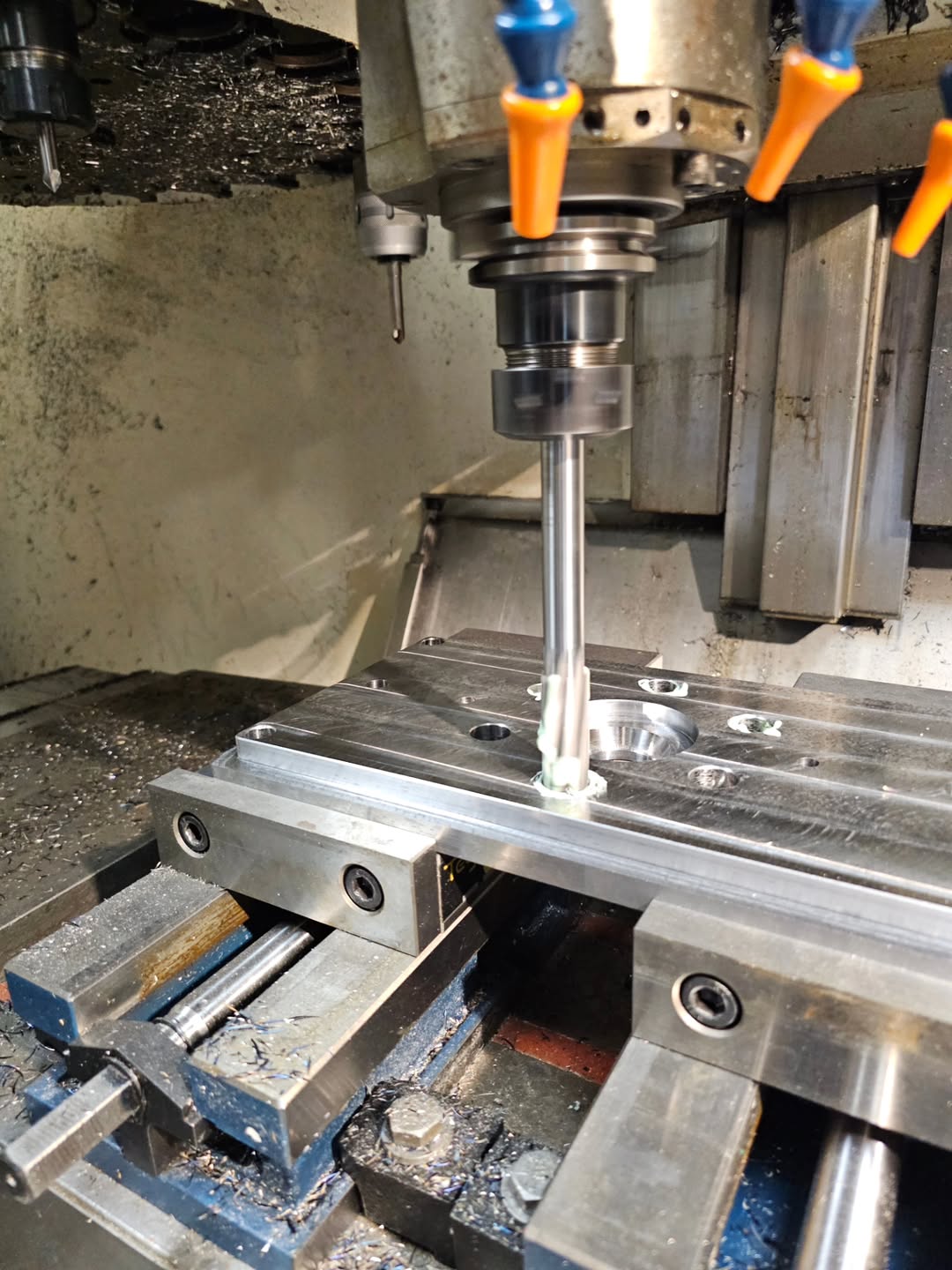 Vertical CNC milling machine cutting a rectangular aluminum plate with a circular pocket and bolt holes, coolant nozzles active