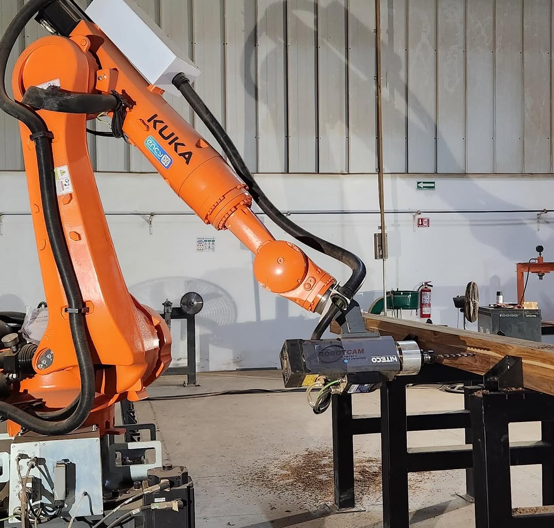 Orange KUKA six-axis industrial robot holding a grinding head against a steel beam in a metal workshop