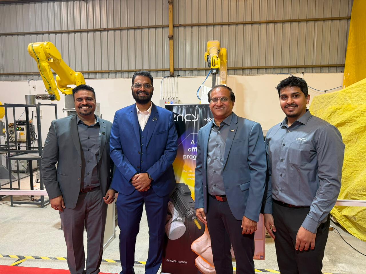 Two yellow 6-axis industrial robots surrounding a demonstration cell with a monitor displaying programming of a curved metal component