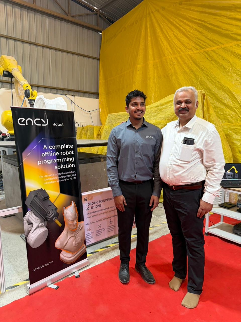 Yellow industrial robot arm positioned behind a sculpted bust display, with a banner and laptop showing robot programming interface, and two men in business attire nearby