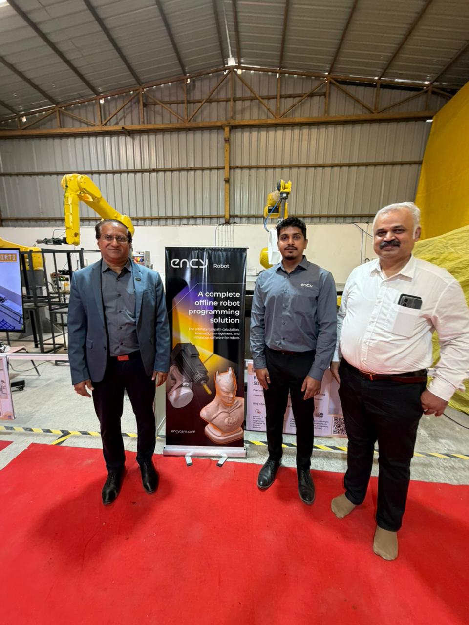 Three men standing in front of an ENCY Robot banner with robotic parts and industrial automation equipment
