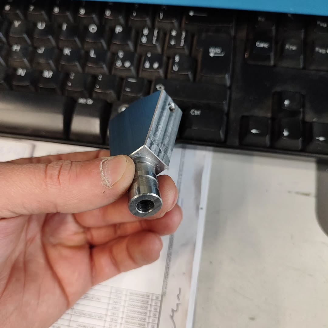 Technician's hand holding a small machined aluminum component with rectangular and cylindrical features, produced on a CNC machine