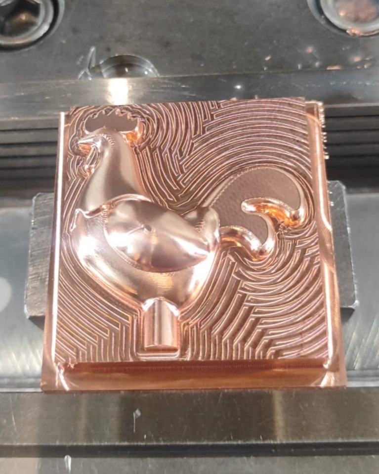 Copper workpiece secured in a metal vise on a CNC milling machine with spiral and parallel tool marks carving a raised bull relief