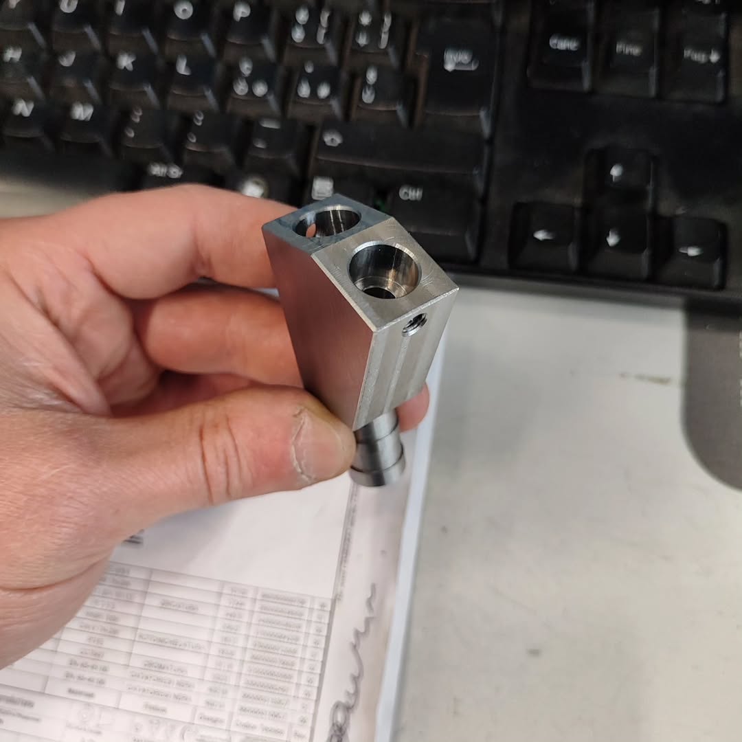Close-up of a machinist's hand holding a small rectangular metal component with precision-milled cylindrical bores and side grooves, produced on a CNC milling machine