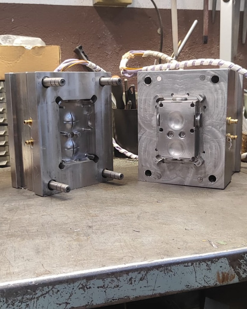 Two precision-machined steel mold halves with twin cavities on a workbench, used for injection molding of plastic components