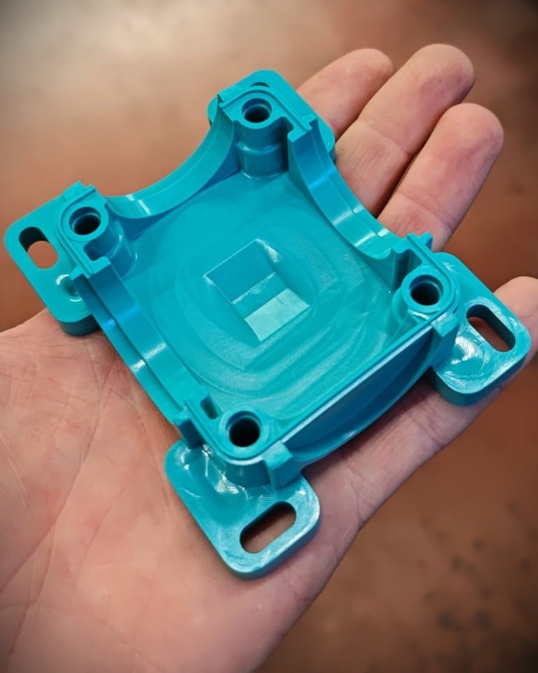 Person's hand holding a small turquoise plastic enclosure with four corner mounting lugs and a recessed central cavity