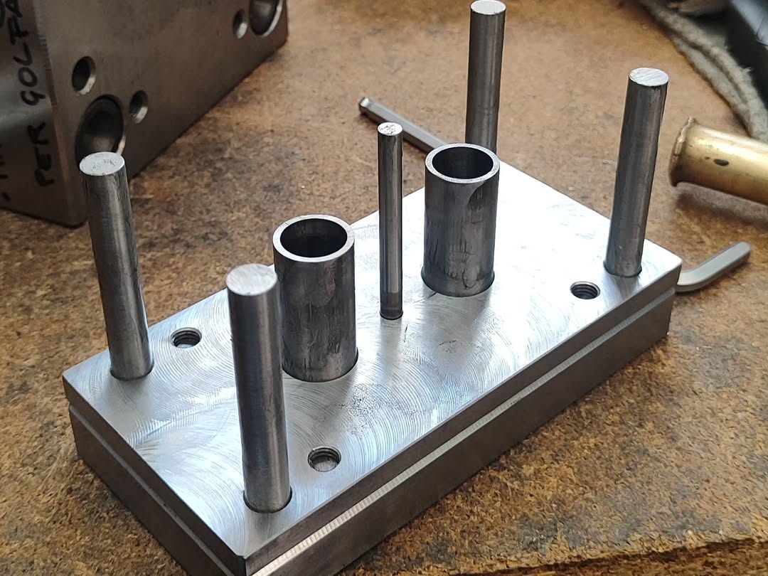 Rectangular steel fixture plate with three hollow bushings and four solid dowel pins on a workbench