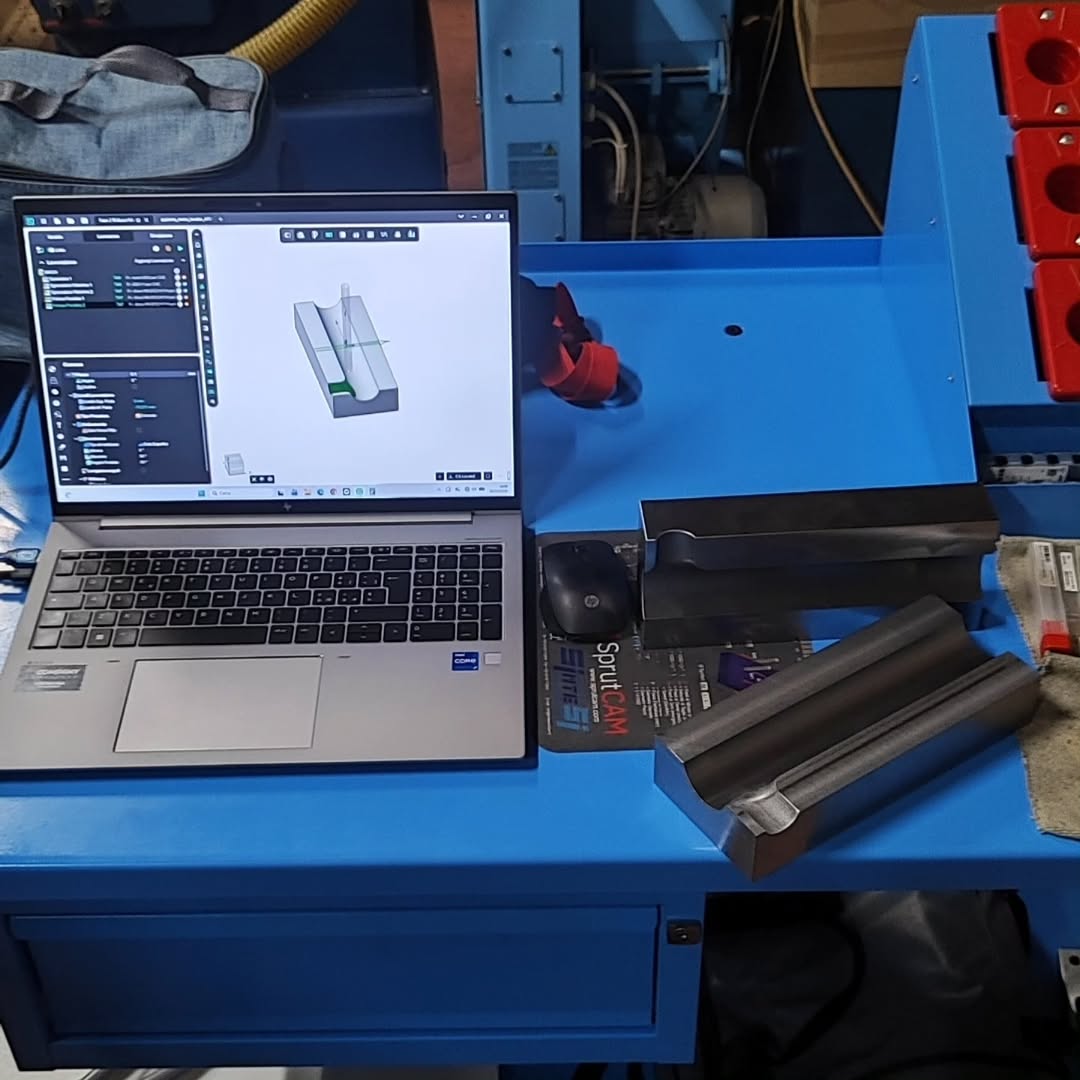 Metal block with dual V-shaped pockets on a blue machine table next to a laptop displaying 3D model and toolpaths in ENCY CAM software