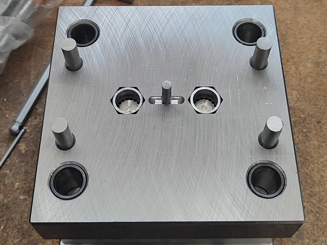 Photograph of a steel mold base with guide pillars, hexagonal cavities, and a central ejector pin, used in plastic or metal tooling