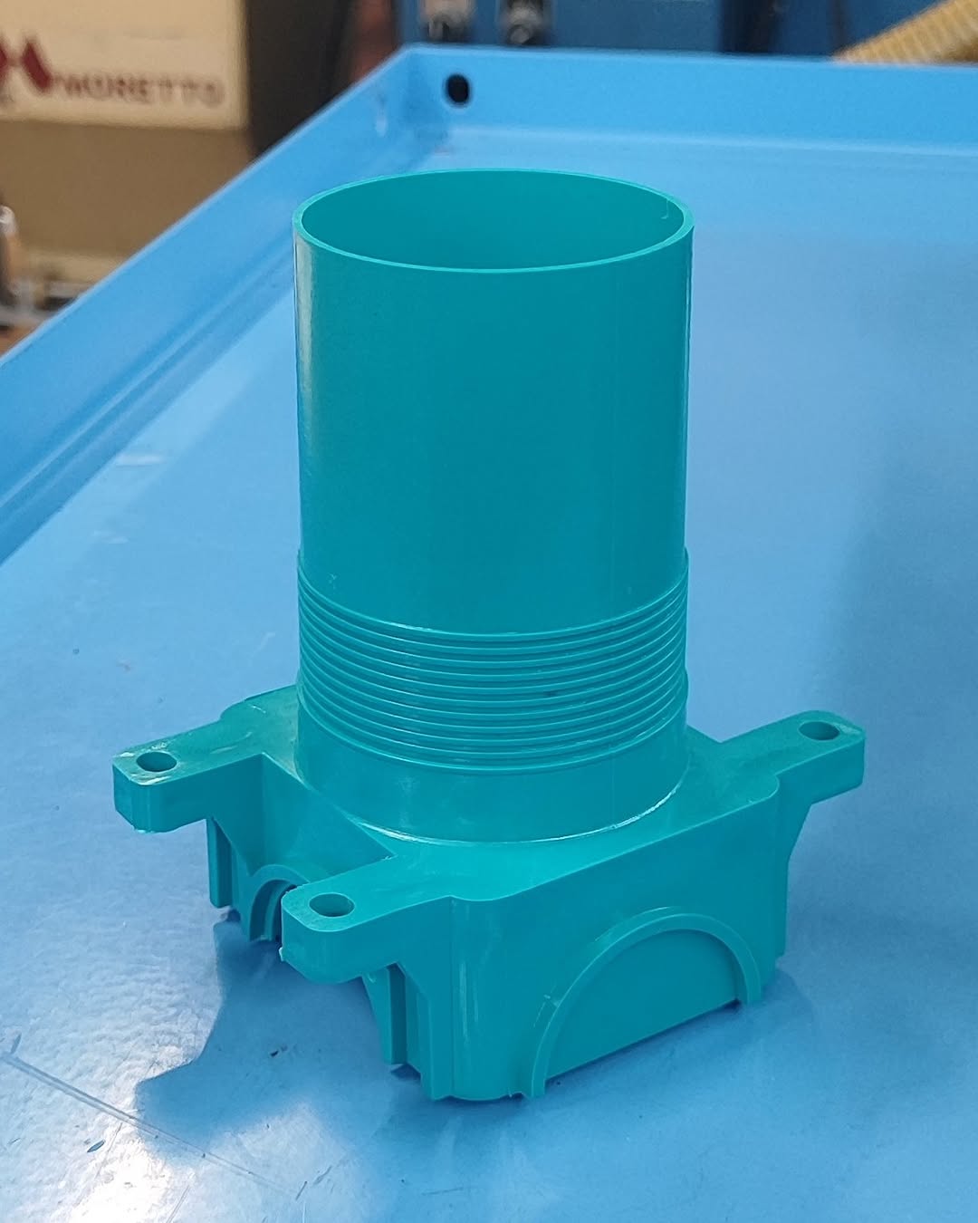Teal cylindrical plastic component with external threads and four mounting flanges on a workbench
