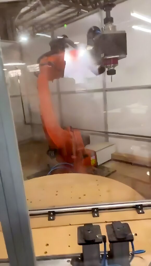 Orange six-axis industrial robot arm with CNC spindle head over a circular fixture table inside a transparent safety enclosure