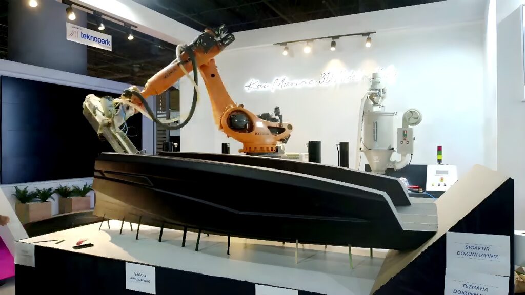 Orange KUKA six-axis industrial robot arm 3D printing a boat hull section with a pellet-fed extruder in a factory setting