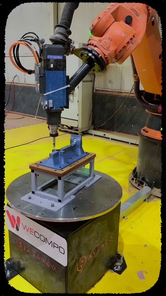 Orange six-axis industrial robot with electric spindle drilling holes in a blue metal casting held on a rotary indexing table in an automated manufacturing cell.