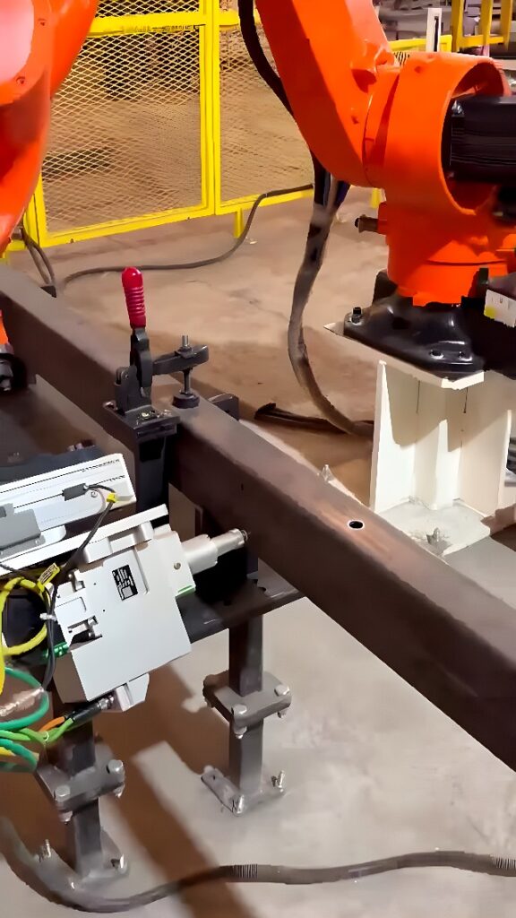 Orange KUKA six-axis robot performing hole-drilling on a clamped rectangular steel tube in an industrial fabrication cell