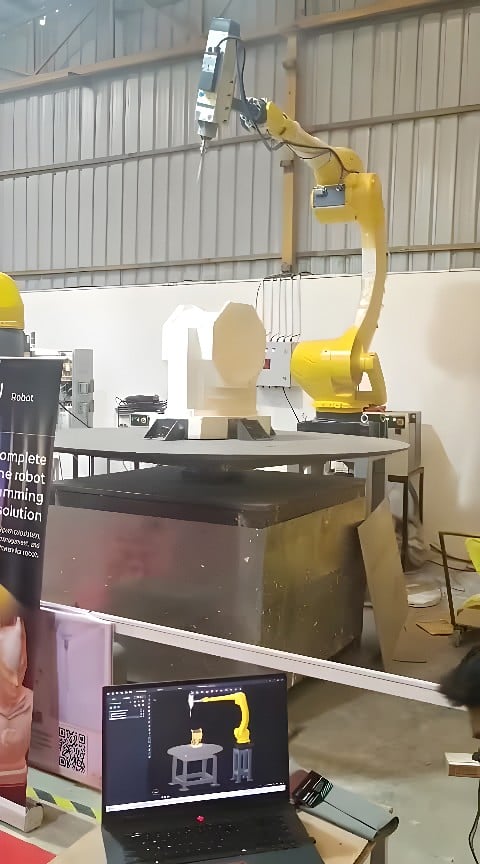 Yellow six-axis industrial robot milling a white polymer block on a rotary table with a laptop showing the toolpath