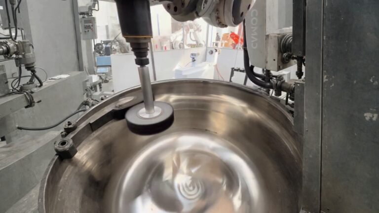 A Comau industrial robot arm performing surface finishing inside a large stainless-steel cylindrical vessel.