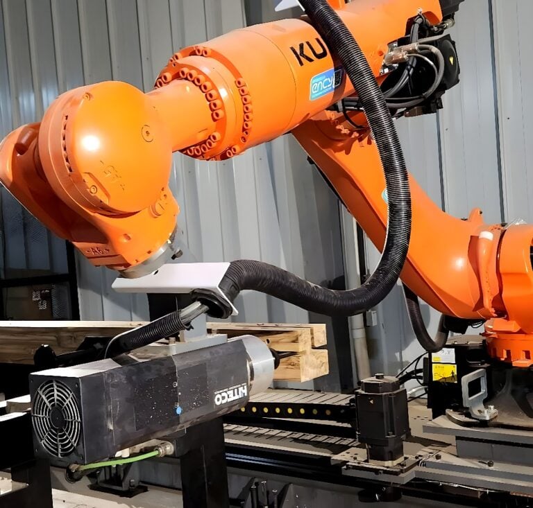 Orange KUKA six-axis industrial robot mounted on linear rails performing automated milling on a metal bracket in a fabrication cell