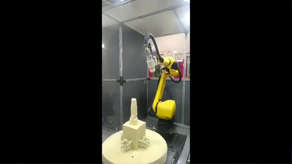 A yellow six-axis industrial robot with a spindle end effector machining a light-colored block inside a safety enclosure.