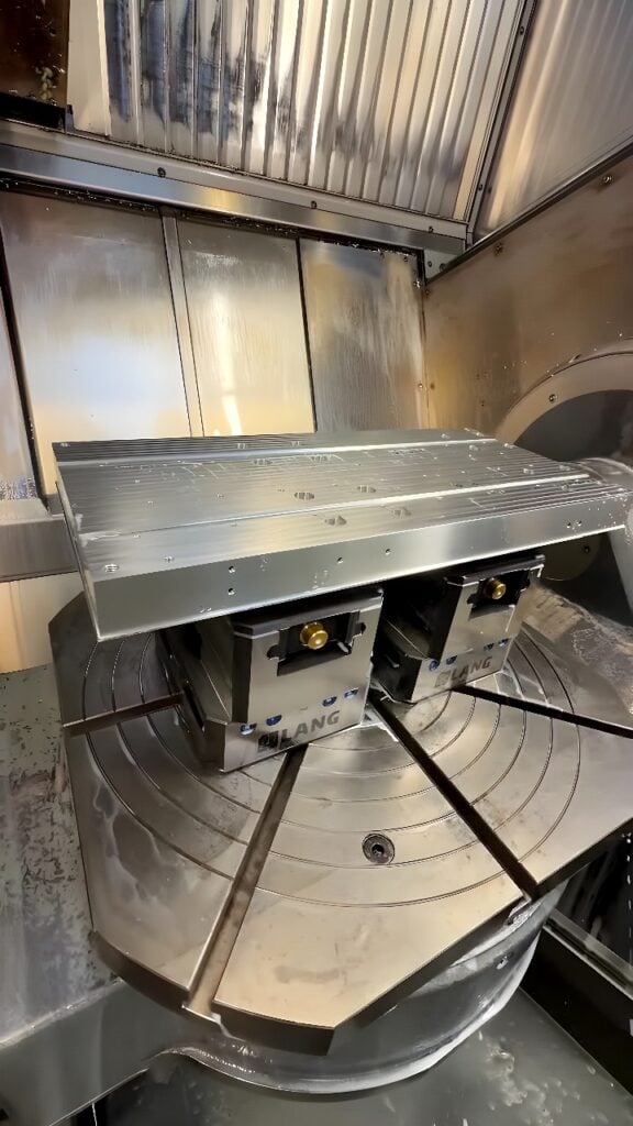 Vertical CNC machining center with a 4th-axis rotary table and dual LANG vises machining an aluminum plate with an end mill, showing parallel slots and counterbored holes.