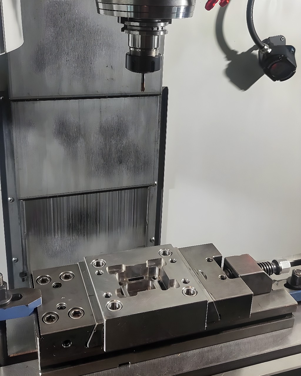 Metal block secured in a vertical CNC milling machine vise with an endmill cutting a central cavity inside an enclosed machining cell