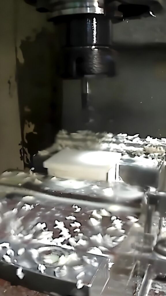 Close-up of a vertical CNC milling machine cutting a white polymer block with an end mill under flood coolant