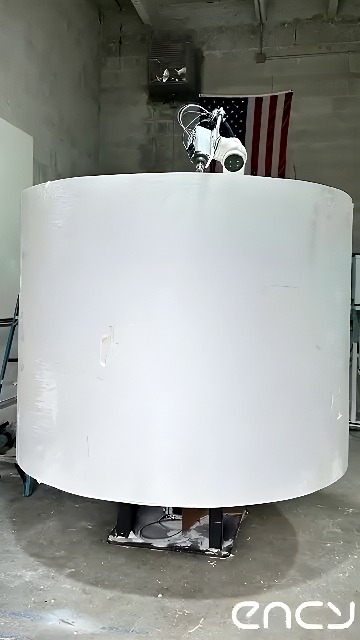 Six-axis industrial robot applying spray coating to a large cylindrical part on a rotary table in a workshop