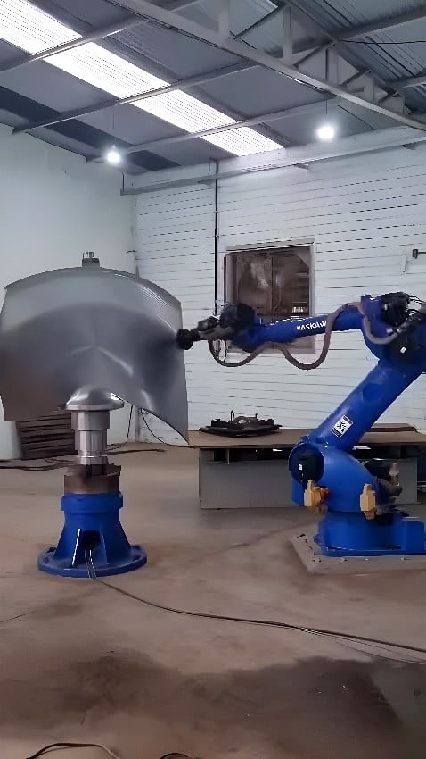 Yaskawa six-axis industrial robot applying an abrasive finishing head to a large curved stainless-steel panel on a powered rotary turntable