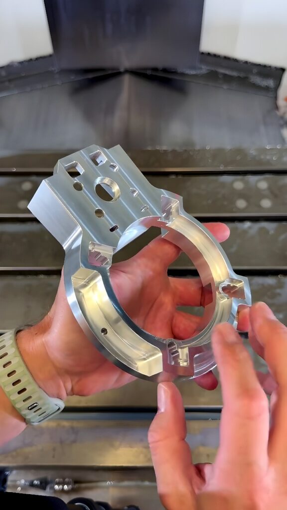 Operator holding a freshly milled aluminum flange-shaped bracket in front of a CNC vertical milling machine