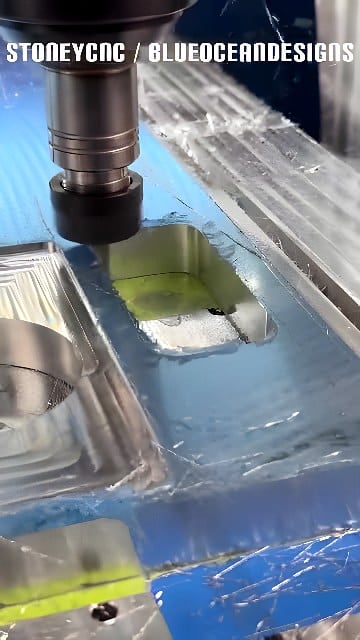 Vertical CNC milling machine cutting a rectangular pocket into a blue-fixtured aluminum workpiece with coolant flowing