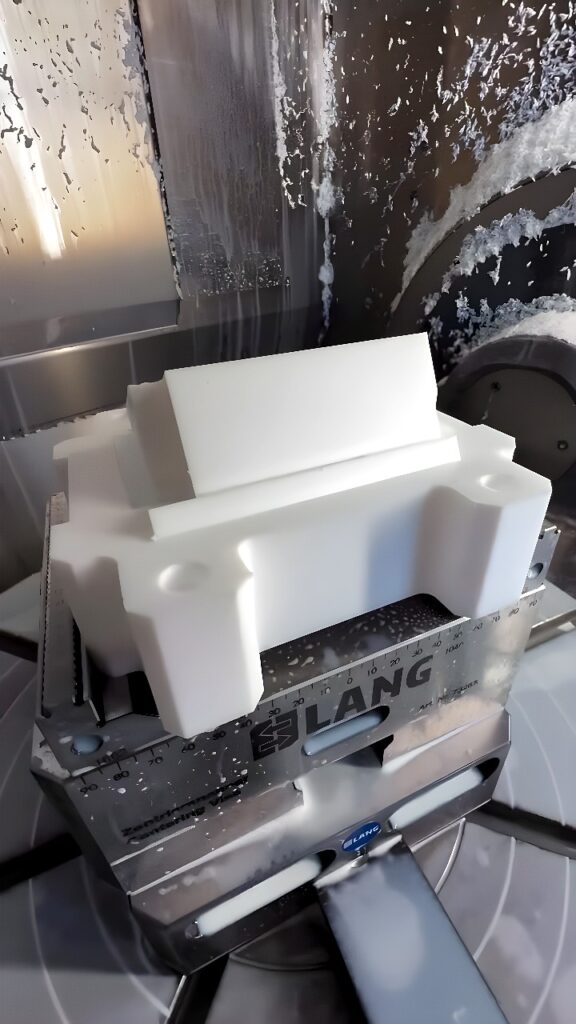 A CNC milling machine cutting a white polymer block held in a Lang modular vise with coolant flowing over the workpiece