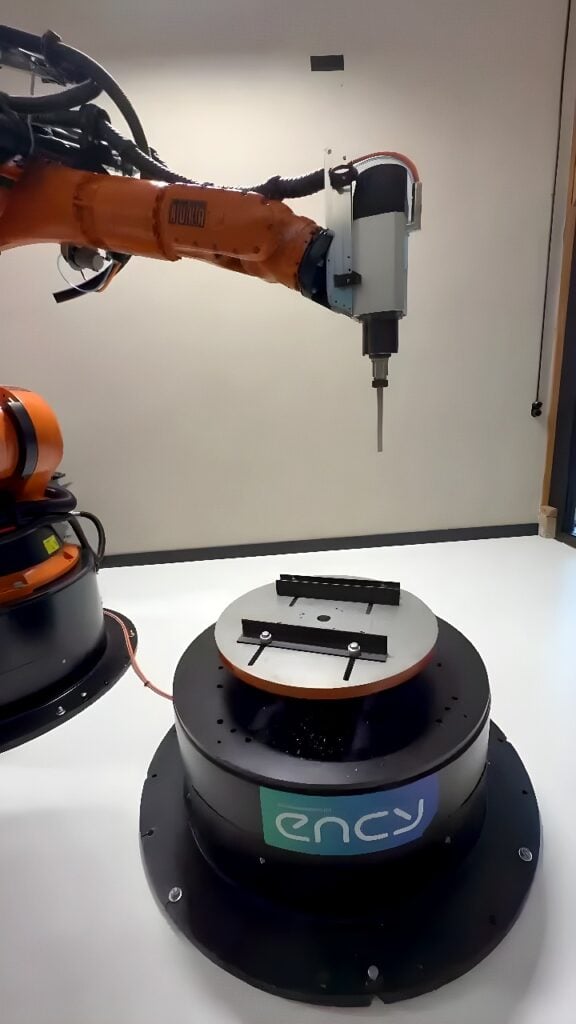 Orange KUKA six-axis industrial robot with a vertical spindle machining a circular aluminum workpiece clamped to a motorized rotary table.