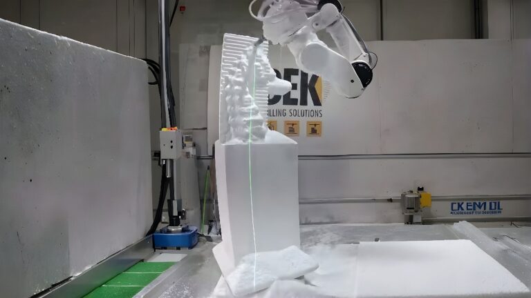 A six-axis industrial robot with a rotating end-mill carves a tall foam block, guided by a green laser line, with foam shavings at the base.