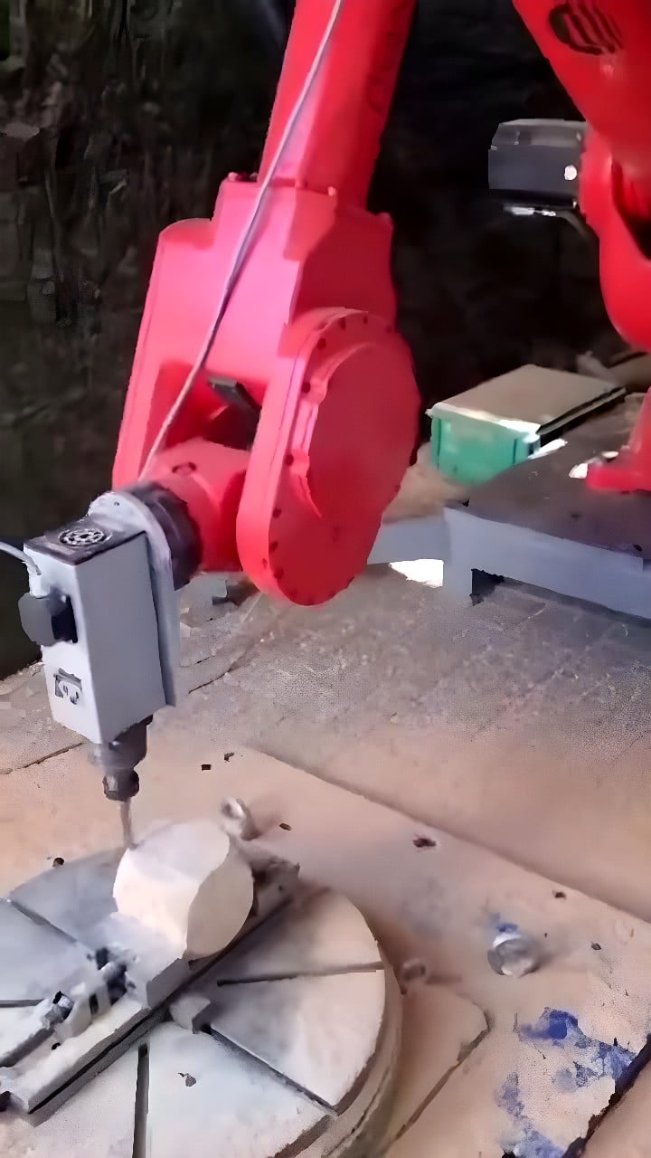 Red six-axis industrial robot with spindle milling head machining a cylindrical blank on a rotary indexing table, producing fine shavings
