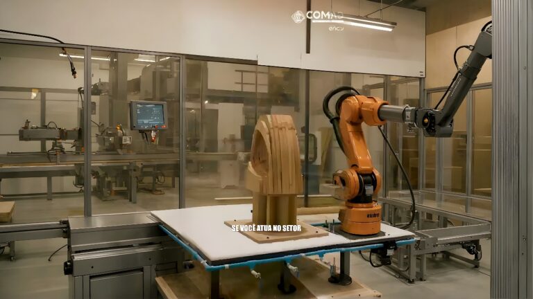 An orange Comau industrial robot arm operates inside a glass-enclosed cell, machining a laminated wood blank on a horizontal table.