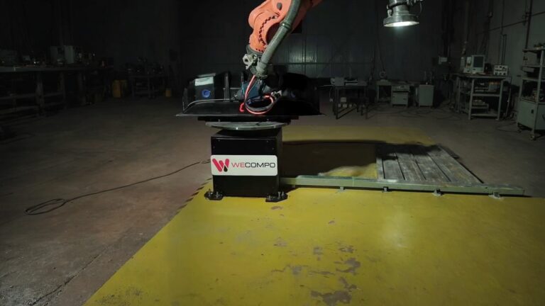 Orange industrial robot arm holding a black molded composite part on a rotary fixture in a manufacturing setting