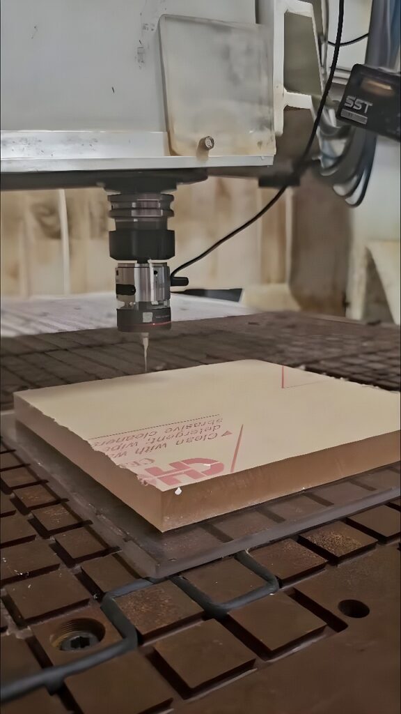 CNC waterjet cutting head positioned above a protective-film-covered acrylic sheet on a metal support table
